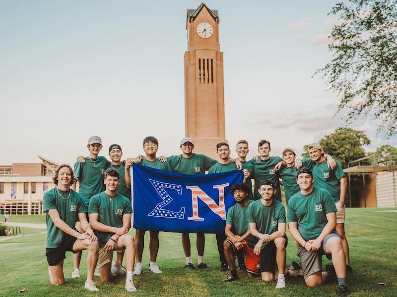 Fraternity members posing in green tees, some kneeling, holding the Sigma Nu flag, in front of the CSU Clock Tower